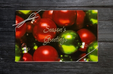 Christmas box covered in red and green ornament design with the words, "Seasons Greetings" embossed in the center. Box is closed with a stretchy silver ribbon. Photo taken on black wooden background.